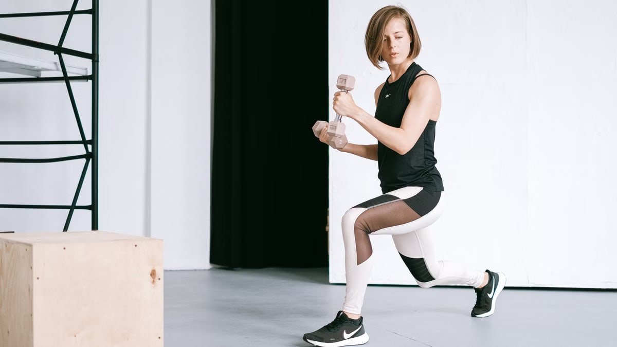 A young woman performing lunges with dumbbells in a modern indoor gym setting.